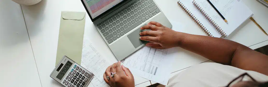 Parcelamento fatura Claro: fotografia com foco nas mãos de uma pessoa que está utilizando o computador. Na mesa, há algumas folhas e calculadora.