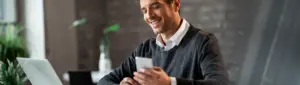 Tipos de investimentos: fotografia de um homem sorrindo enquanto olha para o computador e segura um celular.